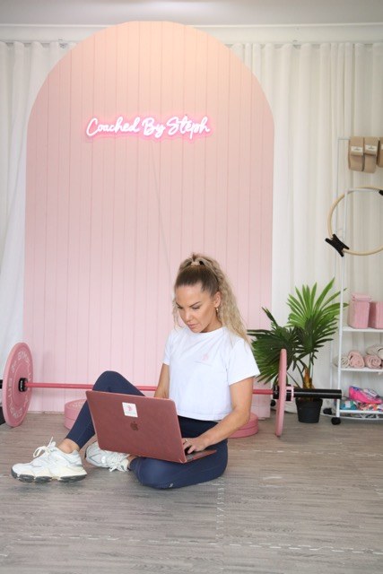 Steph sitting with her laptop beneath the Coached by Steph studio sign